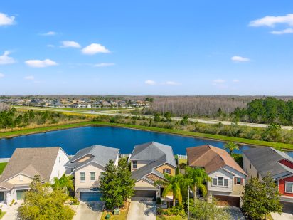 Bright suburban neighborhood with colorful houses, a serene pond, and lush greenery under a clear blue sky.