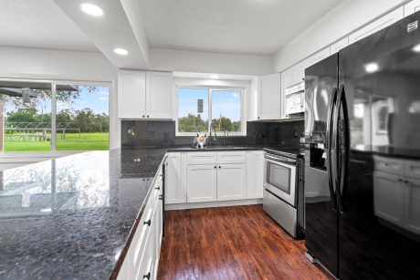 Modern white kitchen with black granite countertops and stainless steel appliances overlooking a green outdoor space.