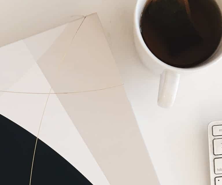 Office desk with a black and white geometric notebook, cup of coffee, and white keyboard, highlighting workspace setup and productivity.