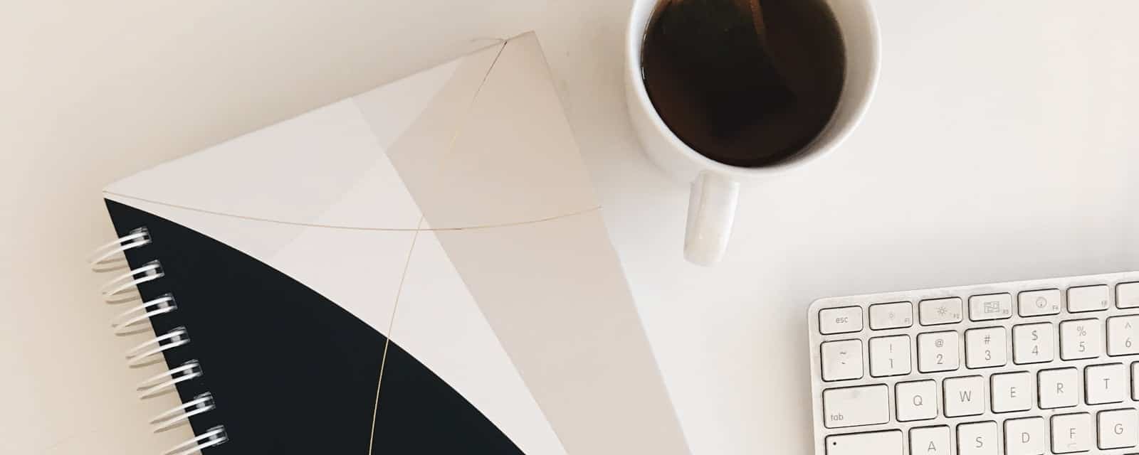 Office desk with a black and white geometric notebook, cup of coffee, and white keyboard, highlighting workspace setup and productivity.